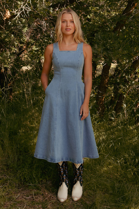 A woman stands on grass among green trees, wearing the Ivy City Co Joanna Dress in Denim and white floral cowboy boots, looking at the camera.