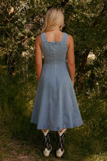 A woman with long blonde hair stands outdoors among tall grass and trees, facing away, wearing the Ivy City Co Joanna Dress in Denim—a sleeveless, light blue midi dress—paired with black and white cowboy boots.