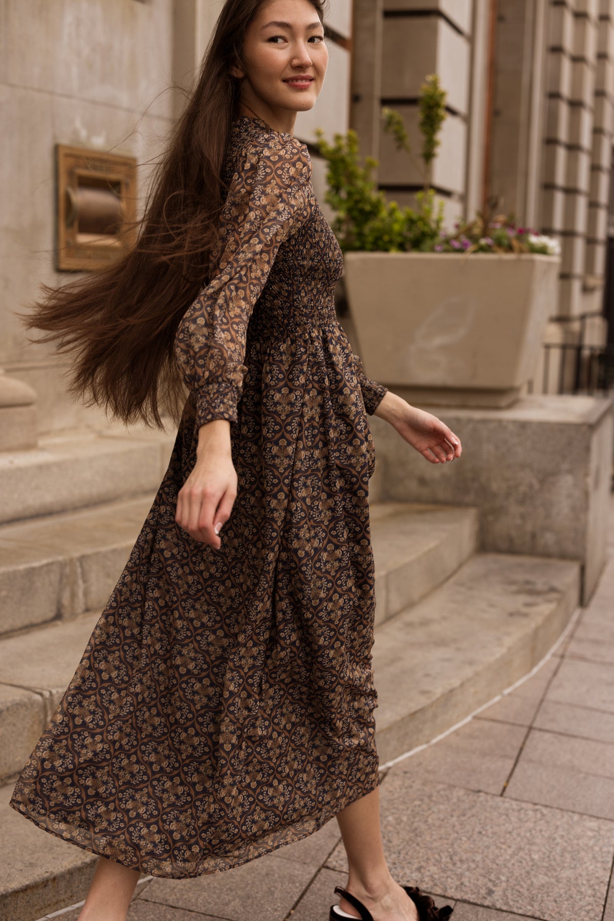 A woman with long hair wearing the Ivy City Co Jordan Dress in Midnight Moroccan Tile walks up stone steps outside a building, smiling slightly.
