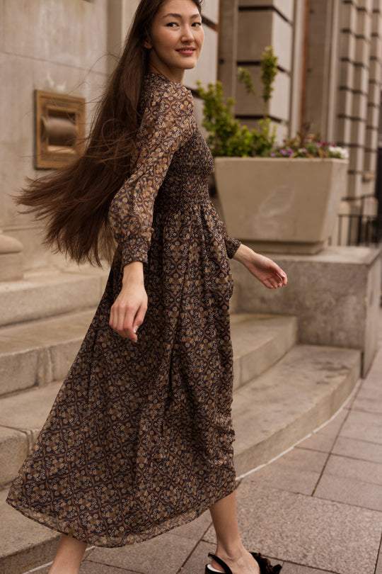A woman with long hair wearing the Ivy City Co Jordan Dress in Midnight Moroccan Tile walks up stone steps outside a building, smiling slightly.