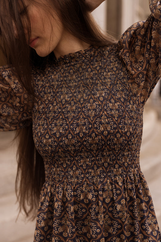 A woman with long brown hair stands outdoors in the Ivy City Co Jordan Dress in Midnight Moroccan Tile, her face partially obscured by her hair and arm, with a softly blurred backdrop of buildings.