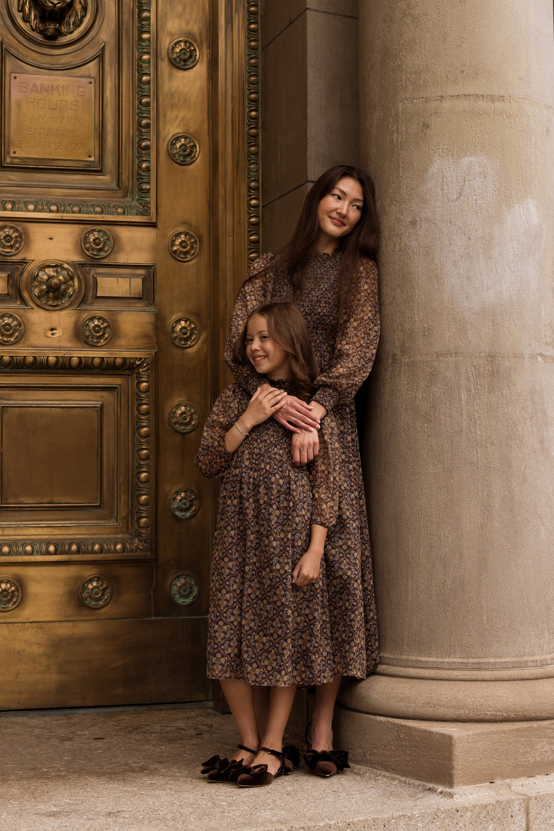 Two women, both dressed in matching Ivy City Co Jordan Dresses in Midnight Moroccan Tile, stand by a large ornate golden door and a stone column, smiling gently as their elegant evening attire complements the setting.