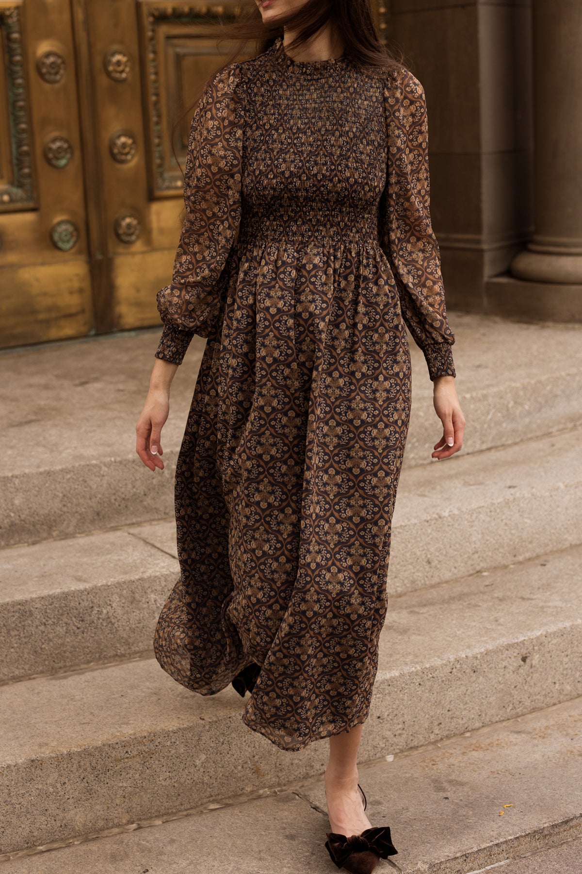 A woman wearing the Ivy City Co Jordan Dress in Midnight Moroccan Tile—a long, sheer maxi dress with intricate patterns and long sleeves—walks down stone steps. Her face is not visible, and ornate gold double doors in the background perfectly capture the elegance of this evening wear.