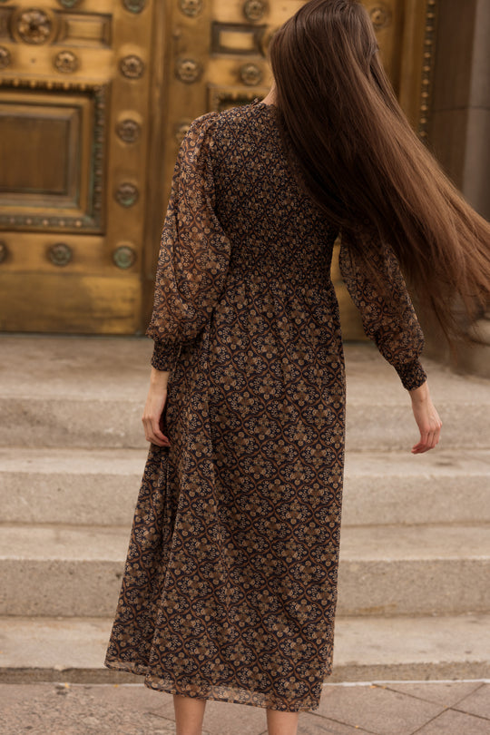 A person with long brown hair wearing the Ivy City Co Jordan Dress in Midnight Moroccan Tile stands on stone steps in front of a large ornate brass door, facing away from the camera.