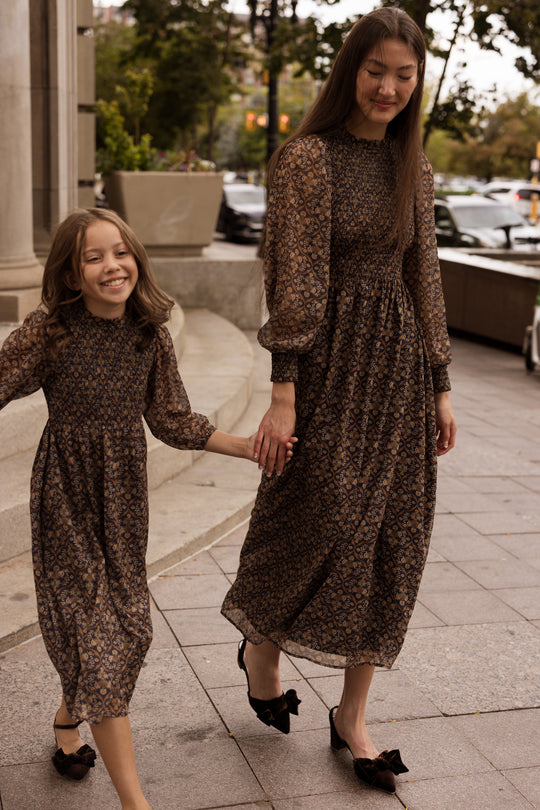 A woman and a young girl, both wearing the Ivy City Co Jordan Dress in Midnight Moroccan Tile paired with black shoes, walk hand in hand on a city sidewalk, smiling and enjoying their time together.