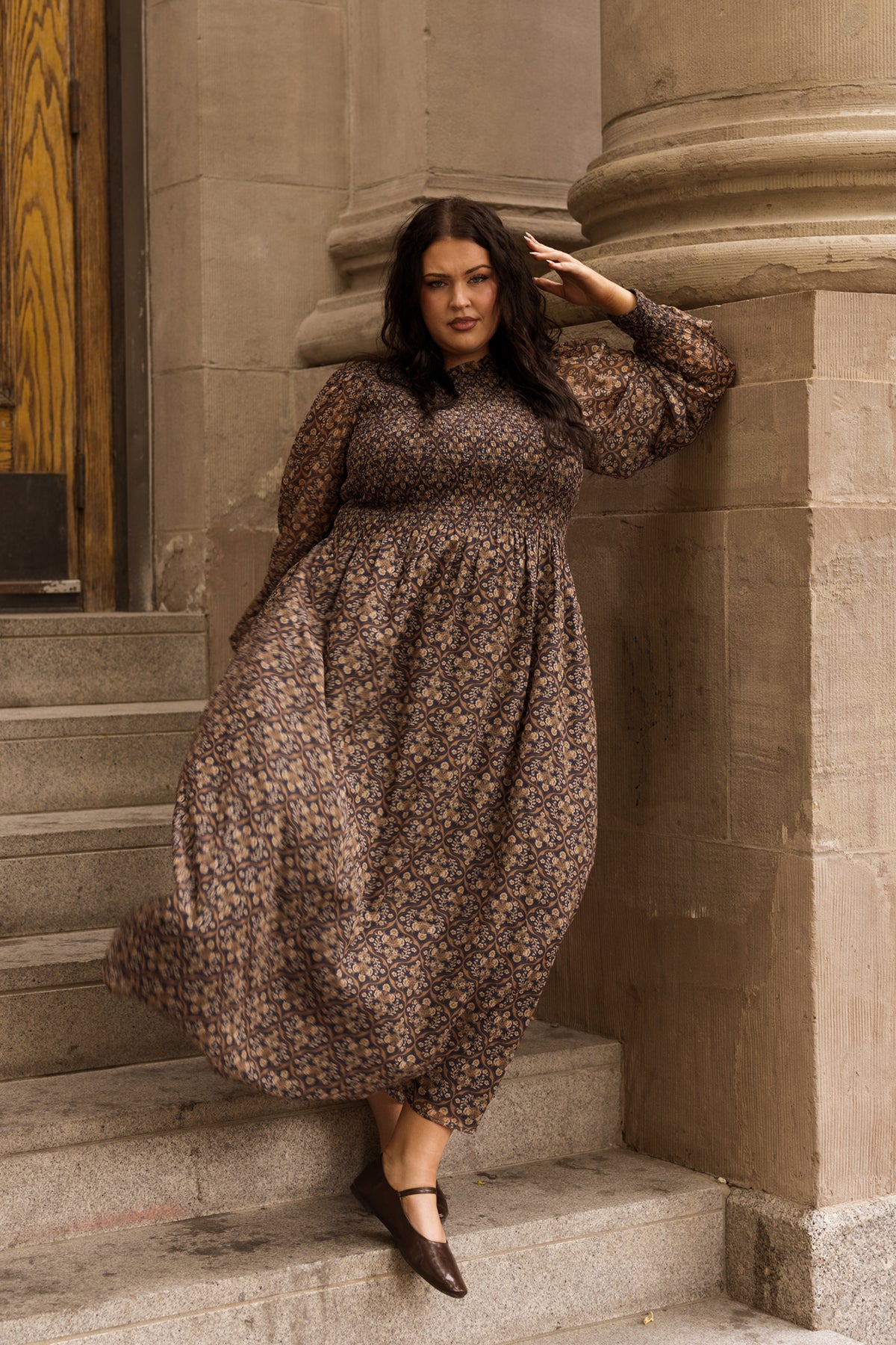 Wearing the Jordan Dress in Midnight Moroccan Tile by Ivy City Co, a woman with loose dark hair leans against a stone column on the steps. One hand rests by her side while the other is raised to her forehead as she looks confidently at the camera.