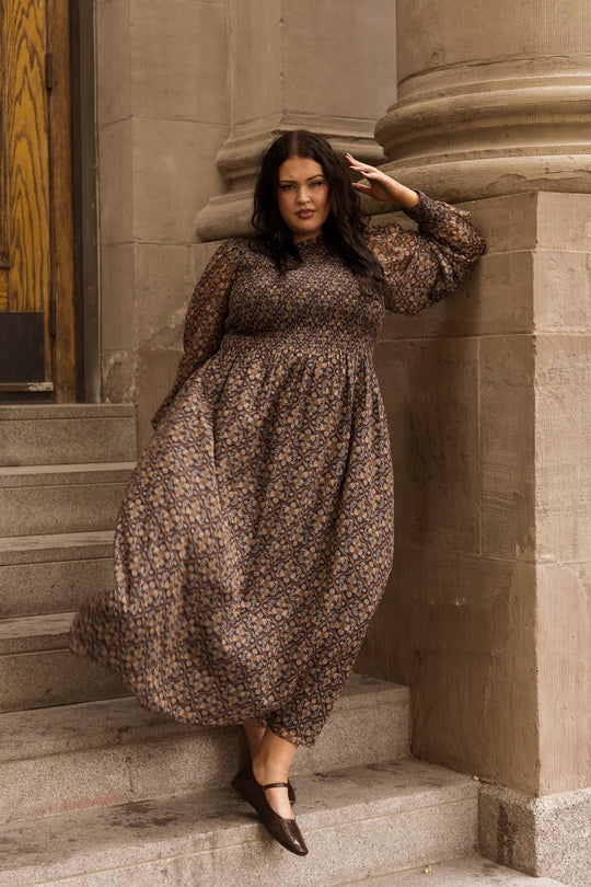 Wearing the Jordan Dress in Midnight Moroccan Tile by Ivy City Co, a woman with loose dark hair leans against a stone column on the steps. One hand rests by her side while the other is raised to her forehead as she looks confidently at the camera.