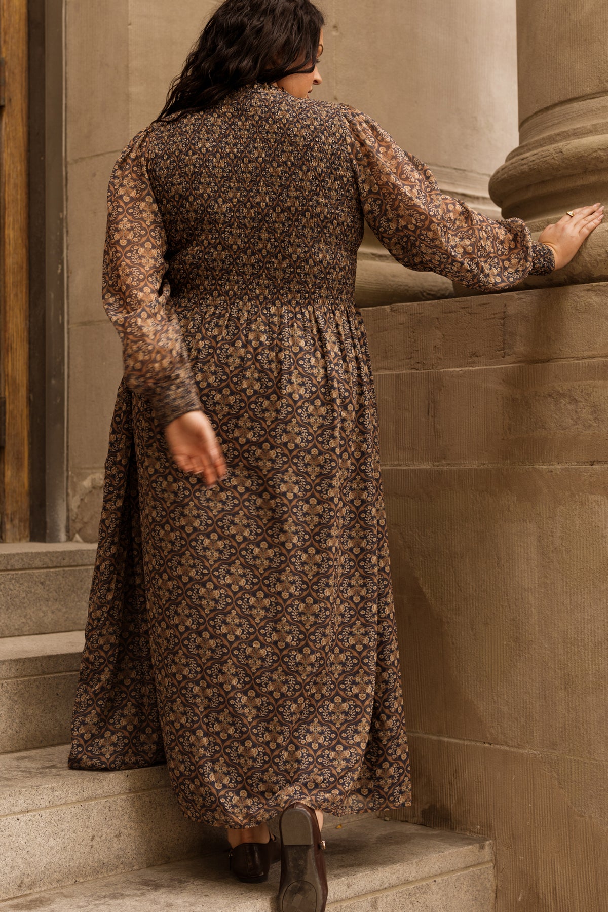 Wearing the Jordan Dress in Midnight Moroccan Tile by Ivy City Co, a person with their back to the camera walks up stone steps, one hand resting on a large column. The long, patterned maxi dress features sheer sleeves and stands out against the textured stone backdrop.