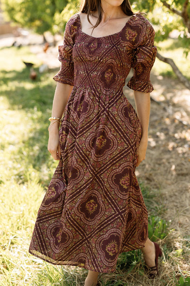 A woman wearing the Josephine Dress in Nocturne Diamond Rose by Ivy City Co, featuring a burgundy floral print with puff sleeves, a smocked bodice, and a square neckline, stands outdoors in a garden setting.