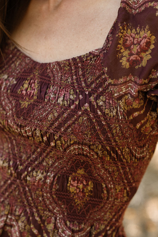 Close-up of a person wearing the Ivy City Co Josephine Dress in Nocturne Diamond Rose, featuring a patterned burgundy fabric with floral designs, smocked bodice details, and a square neckline. The image highlights the texture of the fabric and upper chest area.