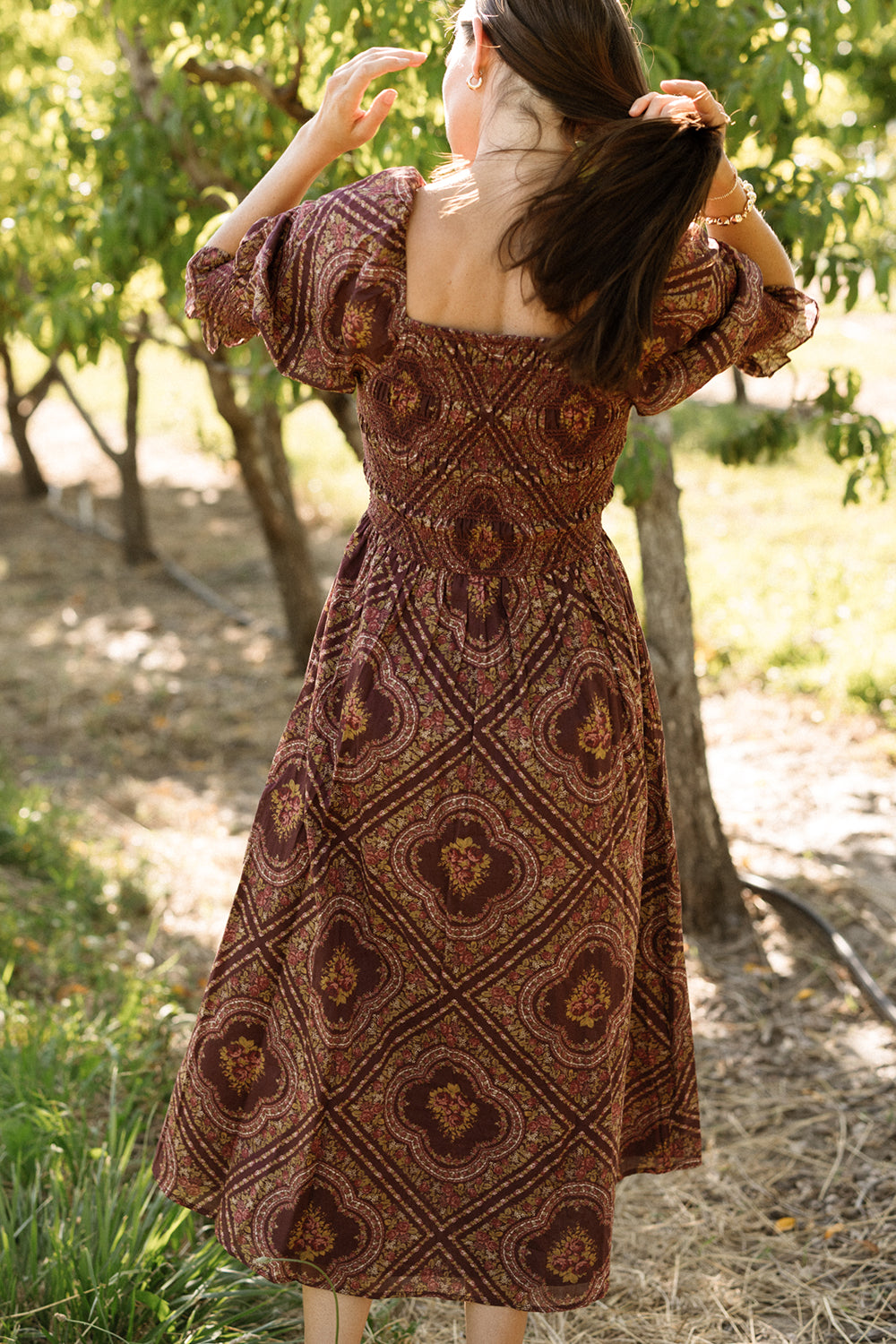 A woman stands outdoors with her back to the camera, holding her hair. She wears the Ivy City Co Josephine Dress in Nocturne Diamond Rose—a brown patterned dress featuring puff sleeves and a smocked bodice—as she stands among trees in a sunlit, grassy area.