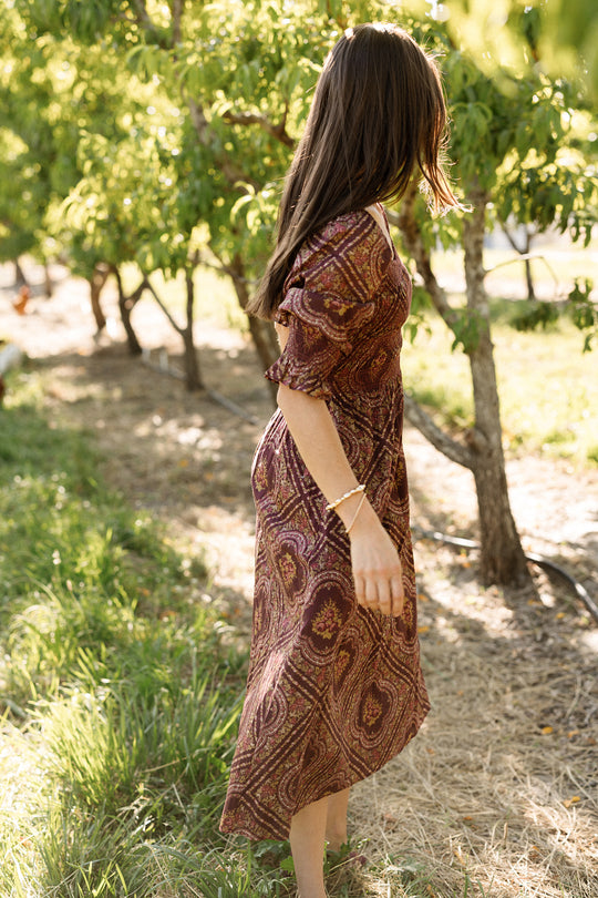 A woman stands barefoot outdoors near green trees, sunlight filtering through the leaves, wearing the Ivy City Co Josephine Dress in Nocturne Diamond Rose. Her hair falls partially over her face as she gazes away from the camera, highlighting the dress’s smocked bodice.