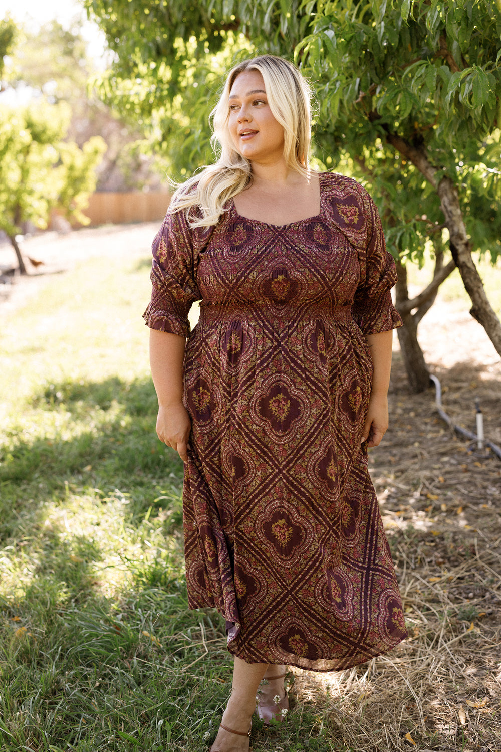 Wearing the Josephine Dress in Nocturne Diamond Rose by Ivy City Co, a woman stands on the grass near lush green trees, smiling softly as sunlight streams through the leaves. The patterned dress features a smocked bodice and long sleeves, enhancing the warm and natural outdoor scene.