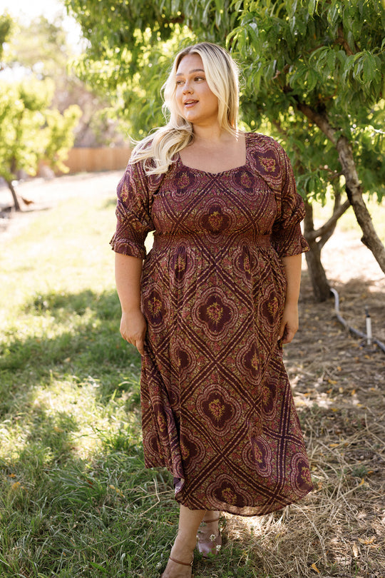 Wearing the Josephine Dress in Nocturne Diamond Rose by Ivy City Co, a woman stands on the grass near lush green trees, smiling softly as sunlight streams through the leaves. The patterned dress features a smocked bodice and long sleeves, enhancing the warm and natural outdoor scene.
