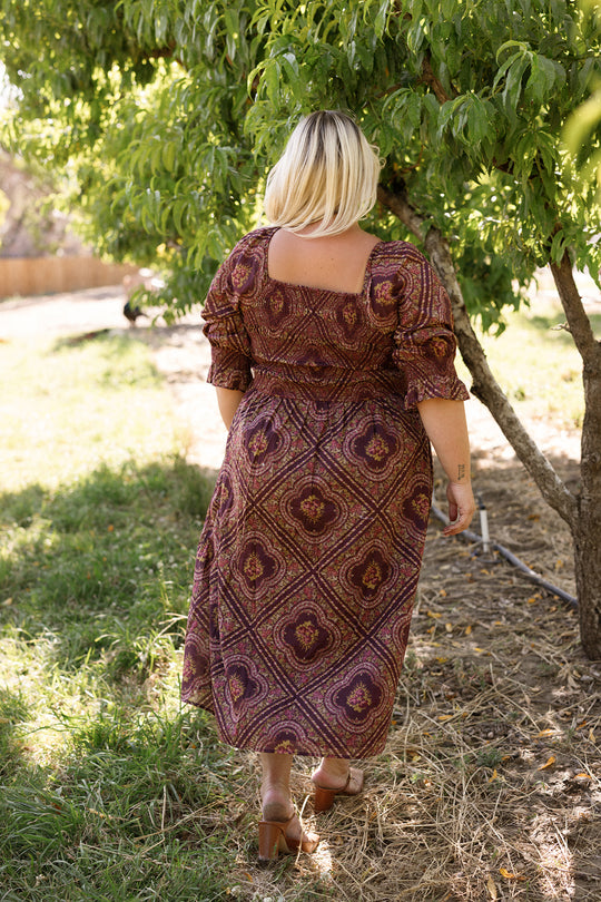 A woman with short blonde hair wearing the Ivy City Co Josephine Dress in Nocturne Diamond Rose walks away on a grassy path beneath a tree, outdoors on a sunny day.