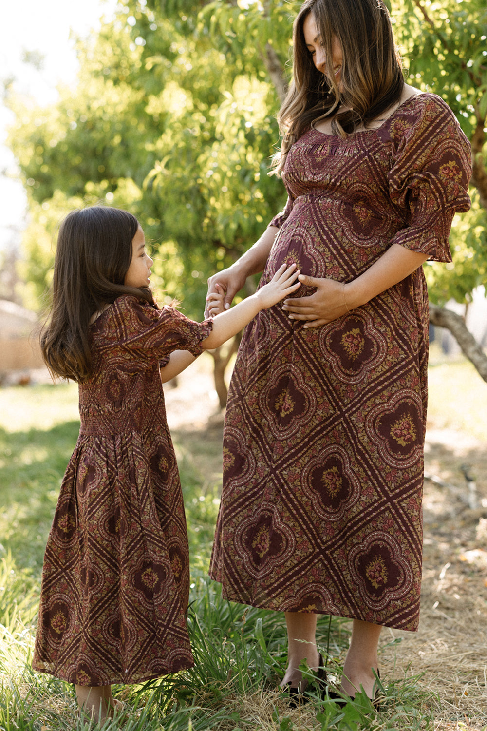 A smiling woman and a young girl, both dressed in matching Ivy City Co Josephine Dresses in Nocturne Diamond Rose, stand outside. The girl touches the woman’s pregnant belly, highlighting her lovely maternity nursing dress. Sunlight filters through green trees behind them.