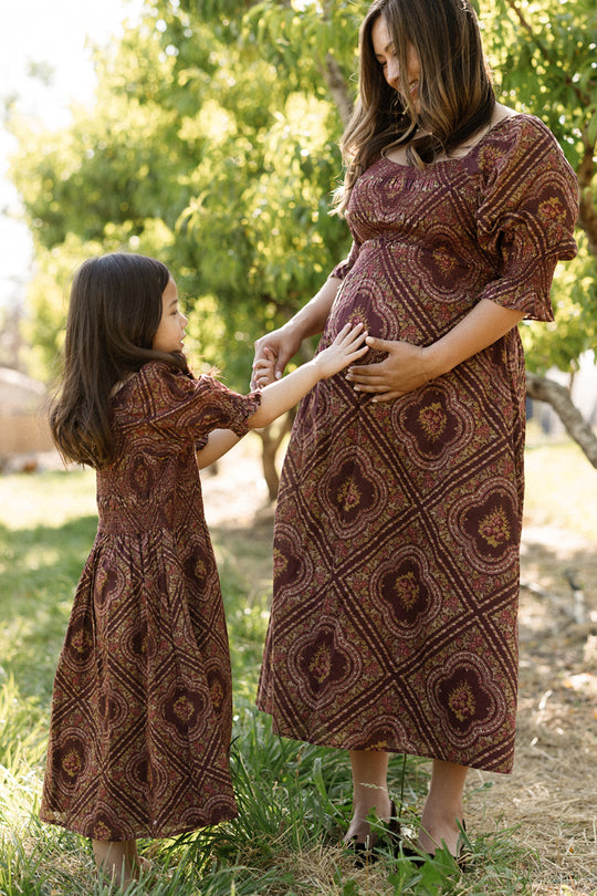 A smiling woman and a young girl, both dressed in matching Ivy City Co Josephine Dresses in Nocturne Diamond Rose, stand outside. The girl touches the woman’s pregnant belly, highlighting her lovely maternity nursing dress. Sunlight filters through green trees behind them.