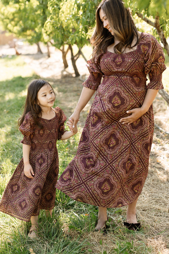 A woman and young girl hold hands outdoors, both wearing matching Ivy City Co Josephine Dresses in Nocturne Diamond Rose with smocked bodices and puff sleeves, standing on grass with trees in the background.