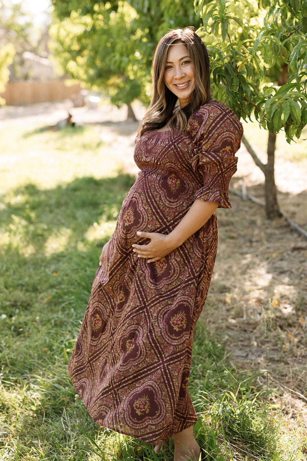 An expectant woman wearing the Ivy City Co Josephine Dress in Nocturne Diamond Rose stands outdoors beside a leafy tree, smiling as she cradles her belly with one hand while sunlight filters through the background trees.