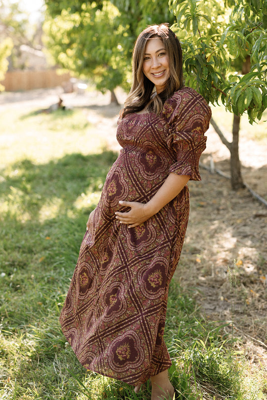 An expectant woman wearing the Ivy City Co Josephine Dress in Nocturne Diamond Rose stands outdoors beside a leafy tree, smiling as she cradles her belly with one hand while sunlight filters through the background trees.