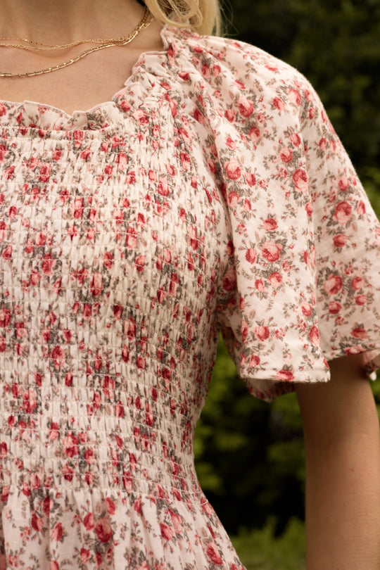 A close-up of a person wearing the Ivy City Co Jovie Dress in Ivory Heritage Rose, which features a smocked bodice and short puff sleeves. Maternity and nursing friendly, it’s ideal for matching mom and me moments with blurred greenery behind.