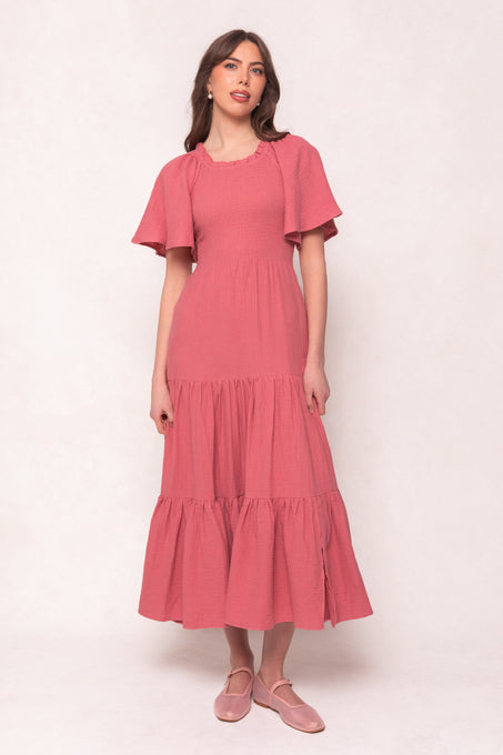 A woman wears the Ivy City Co Jovie Dress in Pink— a pink, short-sleeve, midi-length cotton gauze dress— with pink flats, standing against a plain light background and glancing to the side.