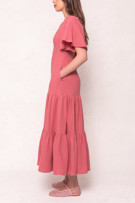 A woman stands in profile wearing the Ivy City Co Jovie Dress in Pink, an ankle-length, tiered cotton gauze maxi with short sleeves and pockets. The stylish, maternity-friendly dress is paired with matching pink flats against a light background.
