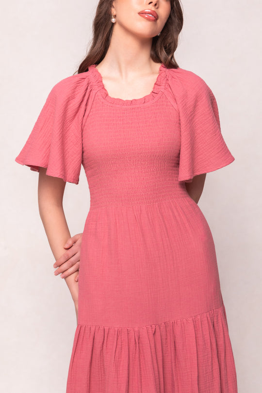 A woman stands with arms loosely crossed against a light background, wearing the Jovie Dress in Pink by Ivy City Co—a textured cotton gauze dress with short sleeves and a ruffled neckline, offering effortless, maternity-friendly style.