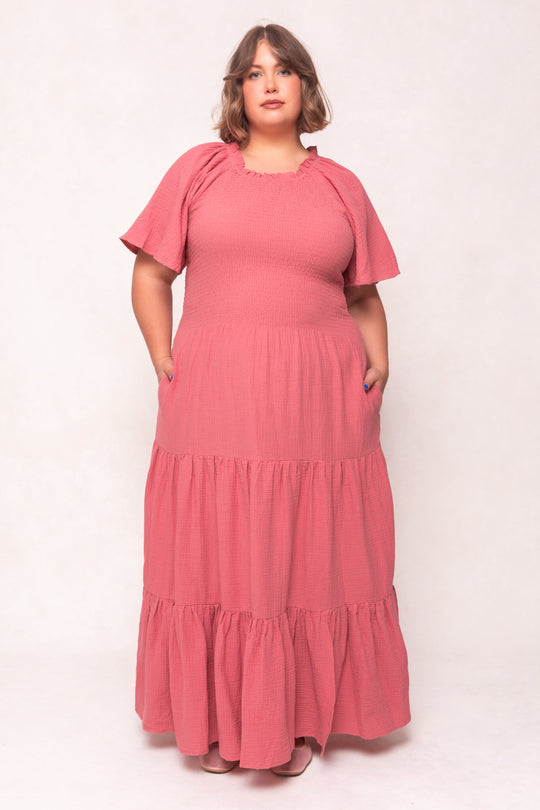 A woman with short, wavy brown hair models the Jovie Dress in Pink by Ivy City Co—a rose-pink cotton gauze maxi with short sleeves and a smocked bodice, maternity friendly—standing confidently against a plain light background.