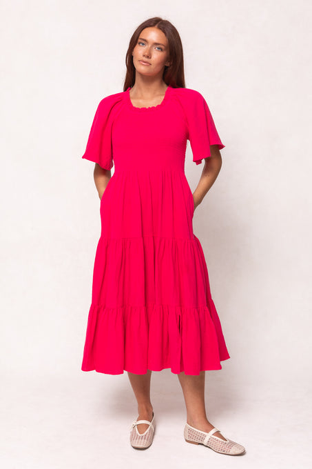 A woman stands against a light background wearing the Ivy City Co Jovie Midi Dress in Hot Pink, a short-sleeved, nursing-friendly cotton dress with smocked bodice, pockets, and neutral flats. Her long brown hair frames her relaxed pose.