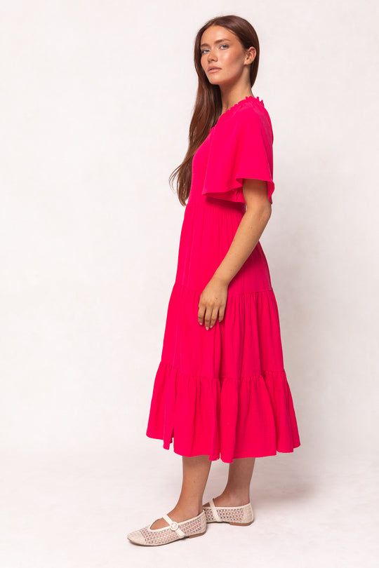 A woman with long brown hair stands sideways against a plain light background, wearing the Ivy City Co Jovie Midi Dress in Hot Pink and white woven flat shoes.