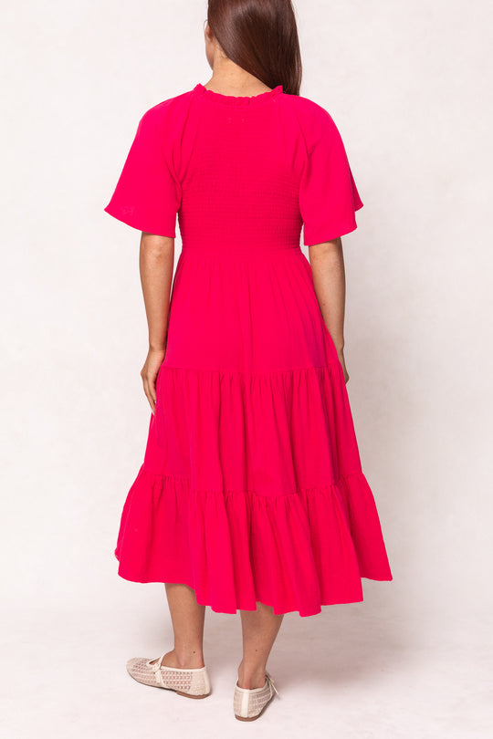 A woman faces away from the camera in the Ivy City Co Jovie Midi Dress in Hot Pink, featuring short sleeves, a smocked bodice, tiered layers, and beige woven slip-ons. Her straight hair is down. A stylish nursing-friendly dress.