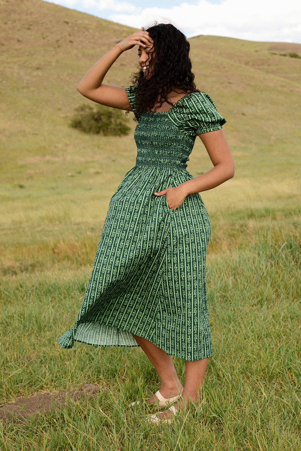 A woman wearing the Ivy City Co Sunday Best Dress in Pine Felicity Floral stands in a grassy field with rolling hills behind her, holding back her curly hair as the wind blows. This dress is both nursing and maternity friendly, offering practicality along with timeless beauty.
