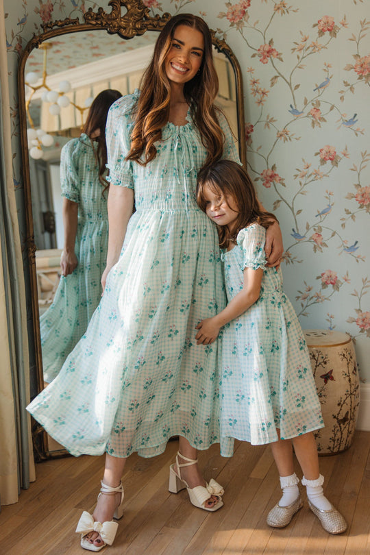 A woman and a young girl, both wearing the Ivy City Co Mini Jessie Dress in Bloom Gingham, smile and hug in front of a mirror in a softly lit, vintage-style room with floral wallpaper.