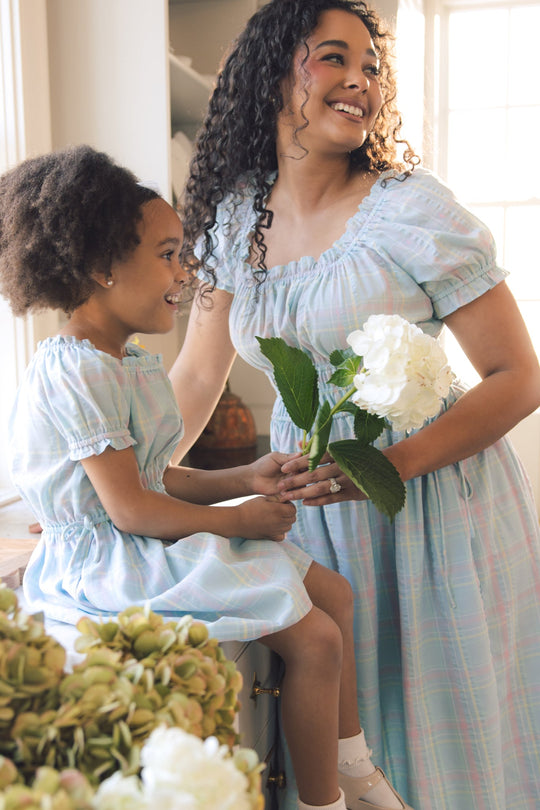 A woman and a girl, both beaming indoors in matching Ivy City Co Thalia Dresses in Pastel Plaid, share a joyful moment. The woman holds a white hydrangea as sunlight streams through the window and fresh flowers in the foreground enhance the bright, charming ambiance of their spring-ready look.