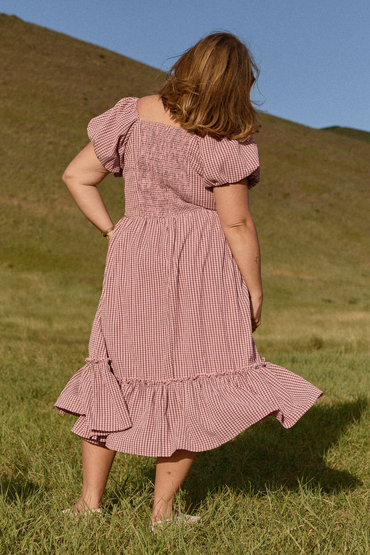 A woman wearing the Ivy City Co Monica Dress in Pink & Brown Gingham stands in a grassy field with hills in the background, facing away from the camera on a sunny day.
