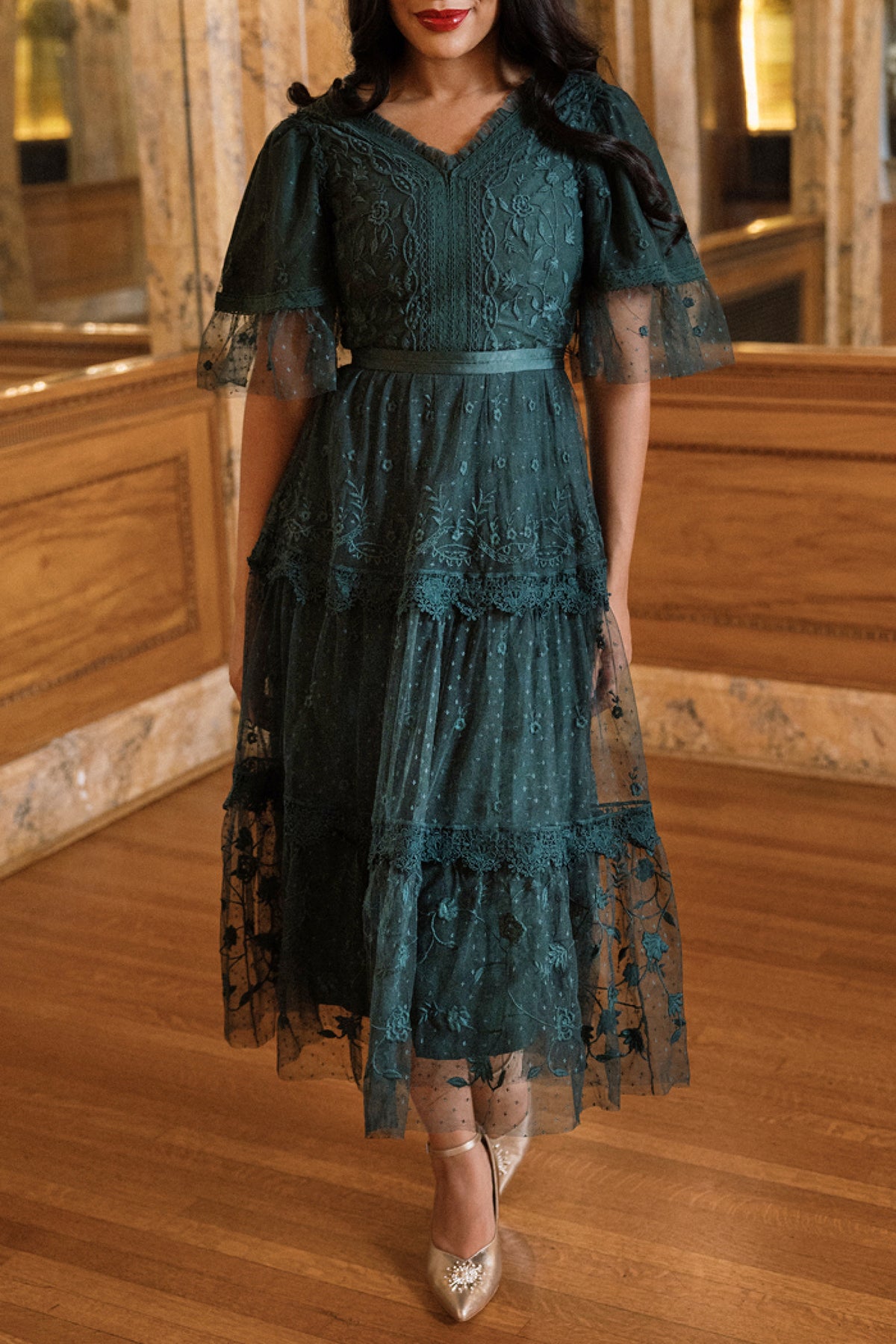 A woman stands indoors on a wooden floor wearing the Ivy City Co Jules Dress in Emerald, which features dark green embroidered lace, tiered layers, and flutter sleeves. She completes the look with metallic pointed heels.