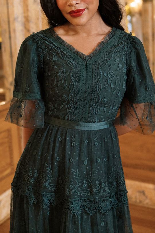 A woman wearing the Jules Dress in Emerald by Ivy City Co—a sophisticated dark green lace dress featuring delicate floral embroidery, sheer ruffled sleeves, and a flattering V-neckline—stands indoors, showcasing this exquisite holiday piece perfect for special occasions.