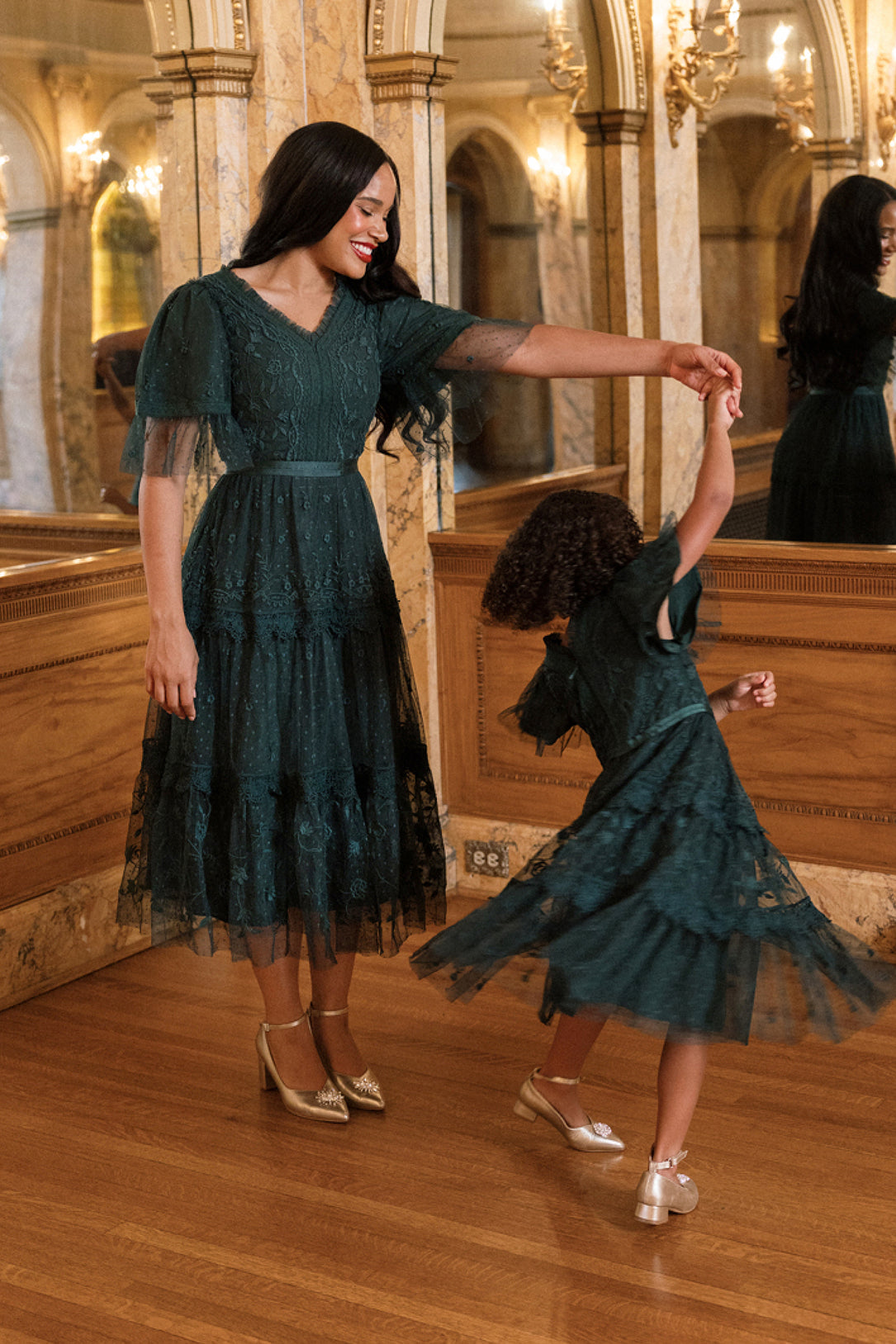 A woman and a young girl, both wearing the Ivy City Co Jules Dress in Emerald, dance together in an elegant, ornate room adorned with mirrors and chandeliers.