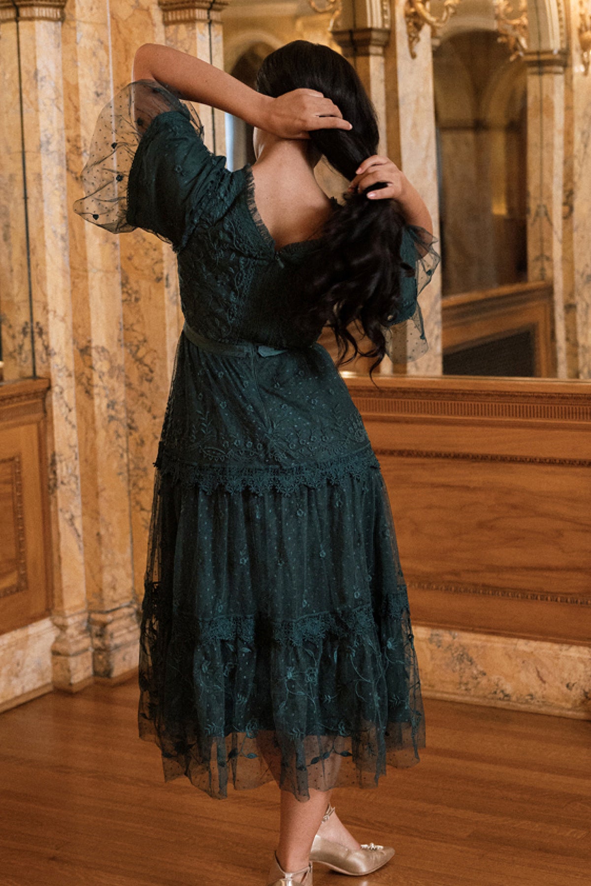 A woman stands indoors on wooden floors, facing away as she adjusts her long dark hair while wearing the Jules Dress in Emerald by Ivy City Co. The dark green embroidered lace of the dress harmonizes with the marble walls and ornate mirrors in the background.