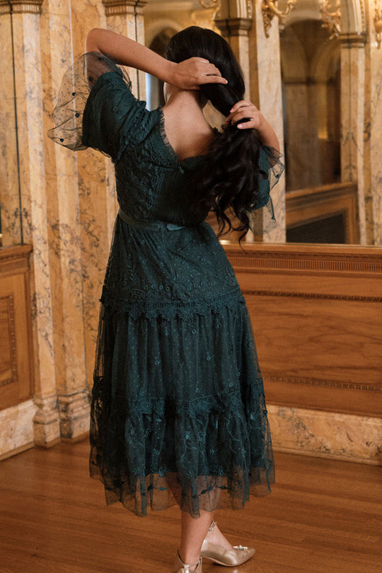 A woman stands indoors on wooden floors, facing away as she adjusts her long dark hair while wearing the Jules Dress in Emerald by Ivy City Co. The dark green embroidered lace of the dress harmonizes with the marble walls and ornate mirrors in the background.