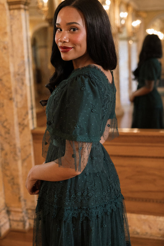 A woman wearing the Jules Dress in Emerald by Ivy City Co stands indoors, smiling and looking over her shoulder. Ornate marble pillars and a mirror reflecting her back can be seen in the background.