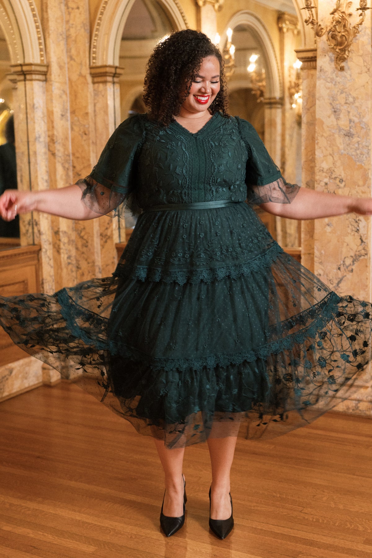 A woman with curly hair twirls joyfully in an elegant, ornate room with mirrored walls and gold decor, wearing the Jules Dress in Emerald by Ivy City Co.