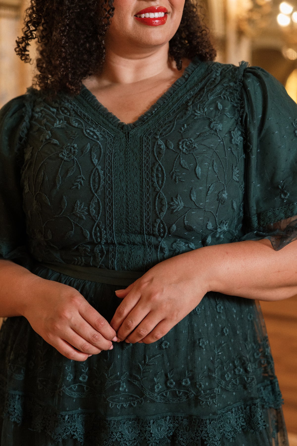 A woman stands indoors with her hands gently clasped in front of her, wearing the Jules Dress in Emerald by Ivy City Co. She has curly hair and red lipstick.