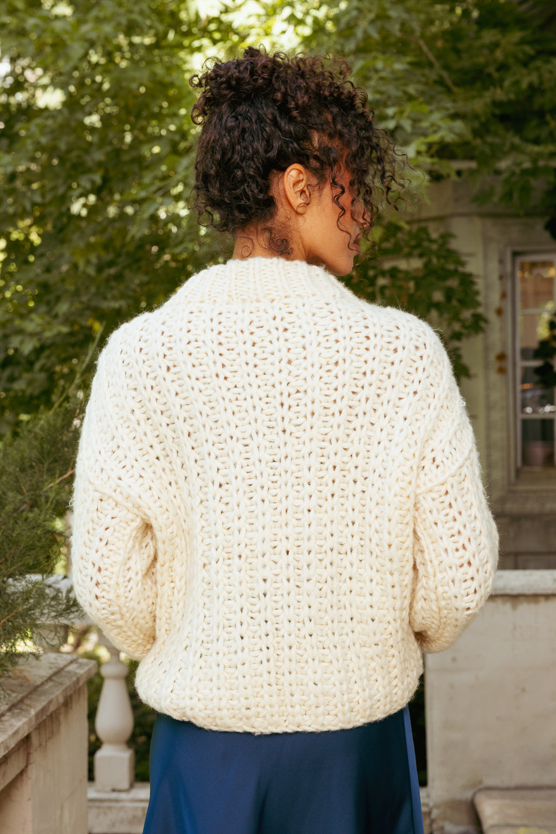 Person wearing a white crochet sweater outdoors with greenery in the background