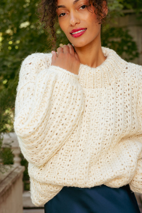 Woman wearing a cream chunky knit sweater with a blurred green outdoor background