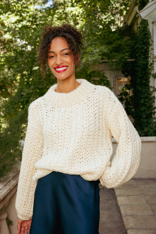 Woman wearing a white sweater and dark blue skirt standing outdoors with greenery in the background