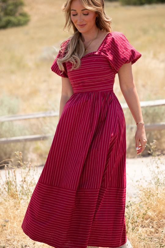 A woman wearing the Julie Dress in Cranberry Pinstripe by Ivy City Co, featuring a smocked bodice and delicate puff sleeves, stands outdoors on a sunny day amidst dry grass and a rustic wooden fence, her loose wavy hair framing a gentle smile.