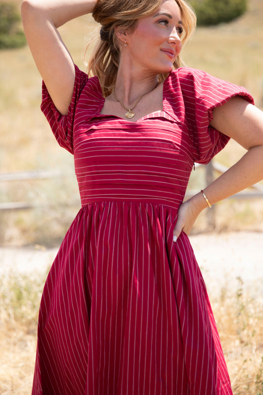 A woman stands outdoors in a sunlit field, wearing the Ivy City Co Julie Dress in Cranberry Pinstripe—a red, short-sleeve dress with a smocked bodice and stripes. She looks to the side, one hand raised to her head and the other on her hip.