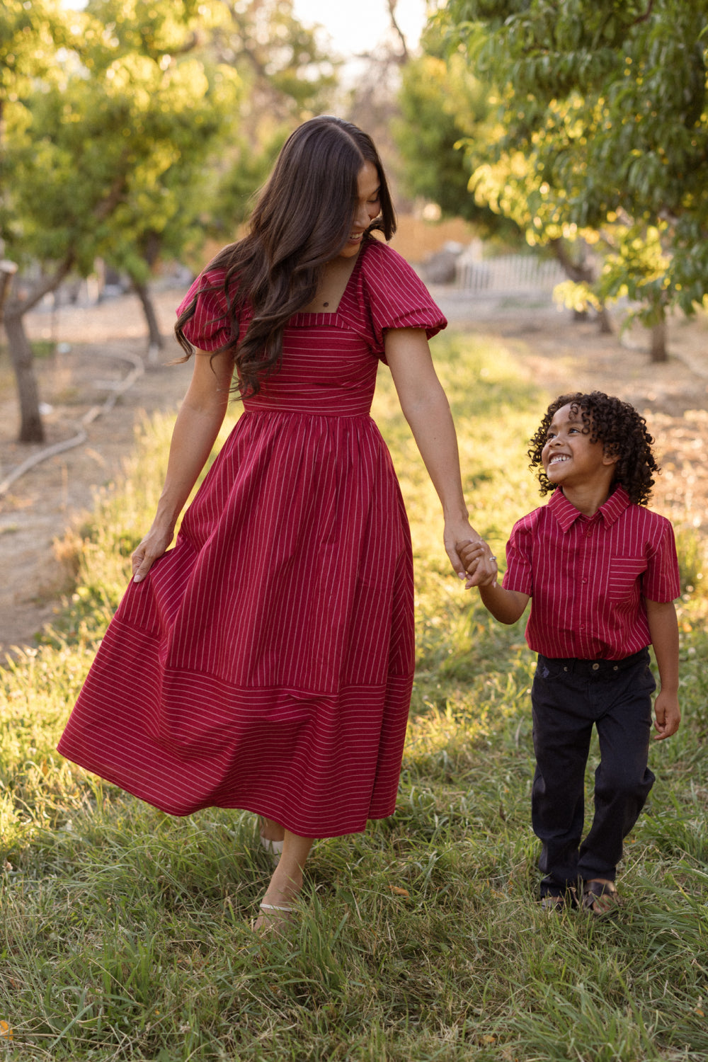 A woman and a young boy, both dressed in red, hold hands and share a smile as they walk together along a grassy path lined with trees. The woman’s Ivy City Co Julie Dress in Cranberry Pinstripe features puff sleeves and a charming smocked bodice, adding an elegant touch to their joyful outing.